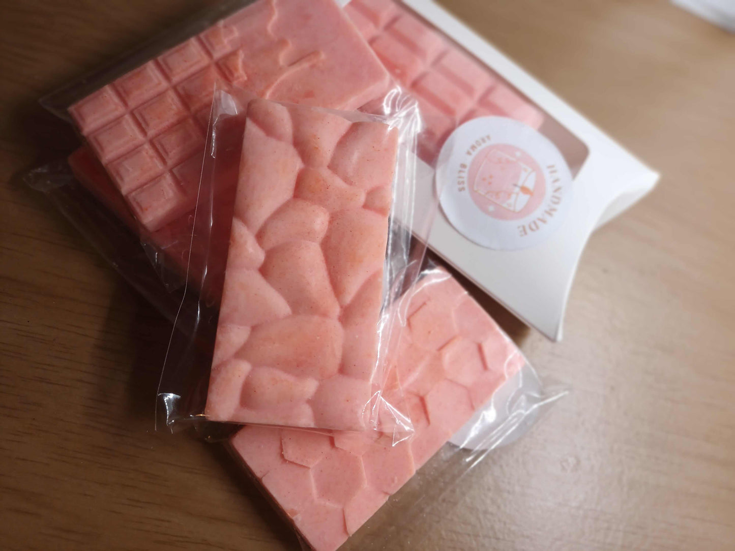 Two pack of pink hand-poured soy wax melts with textured surface in transparent wrappers, displayed on wooden surface with white packaging box.